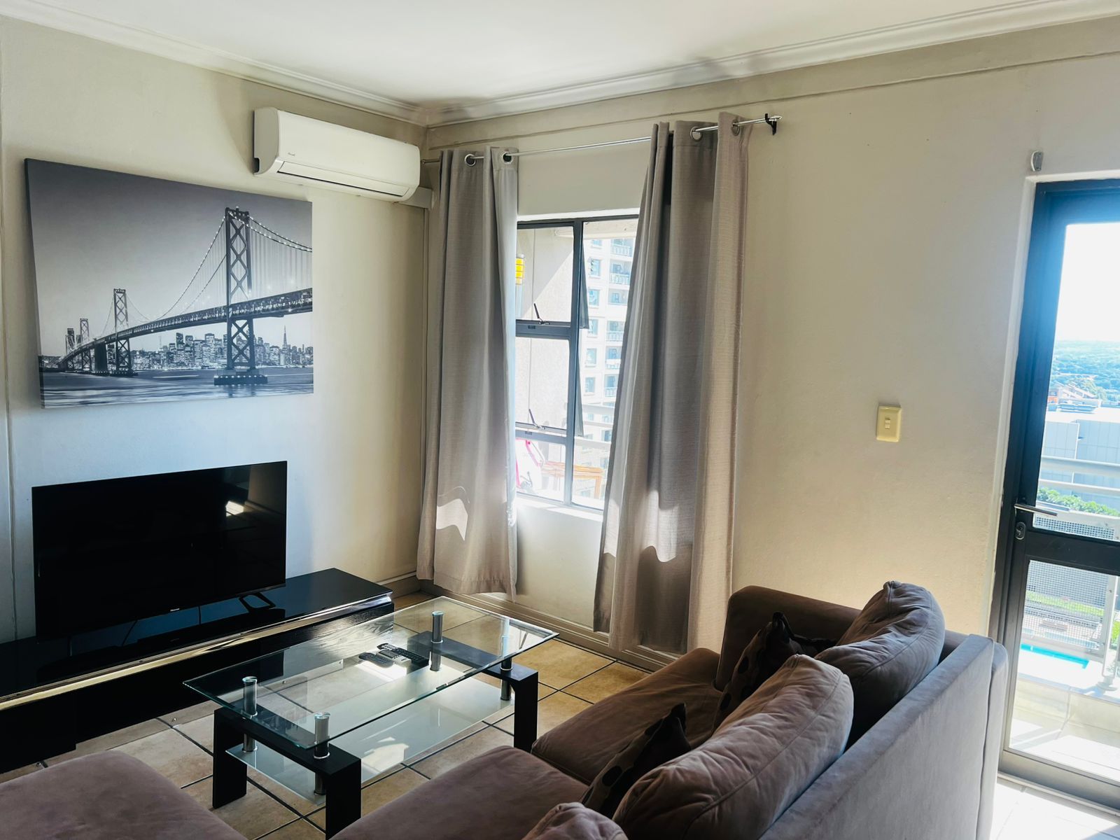 A living room with a flat-screen TV, a glass coffee table, a sofa, and a large window with curtains.