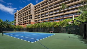 Ein Tennisplatz mit Basketballkorb, umgeben von einem Zaun und Palmen, mit einem mehrstöckigen Hotel im Hintergrund.