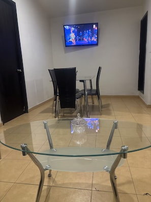 Una habitación con una mesa de comedor de vidrio, sillas negras, un televisor montado en la pared y suelo de baldosas.