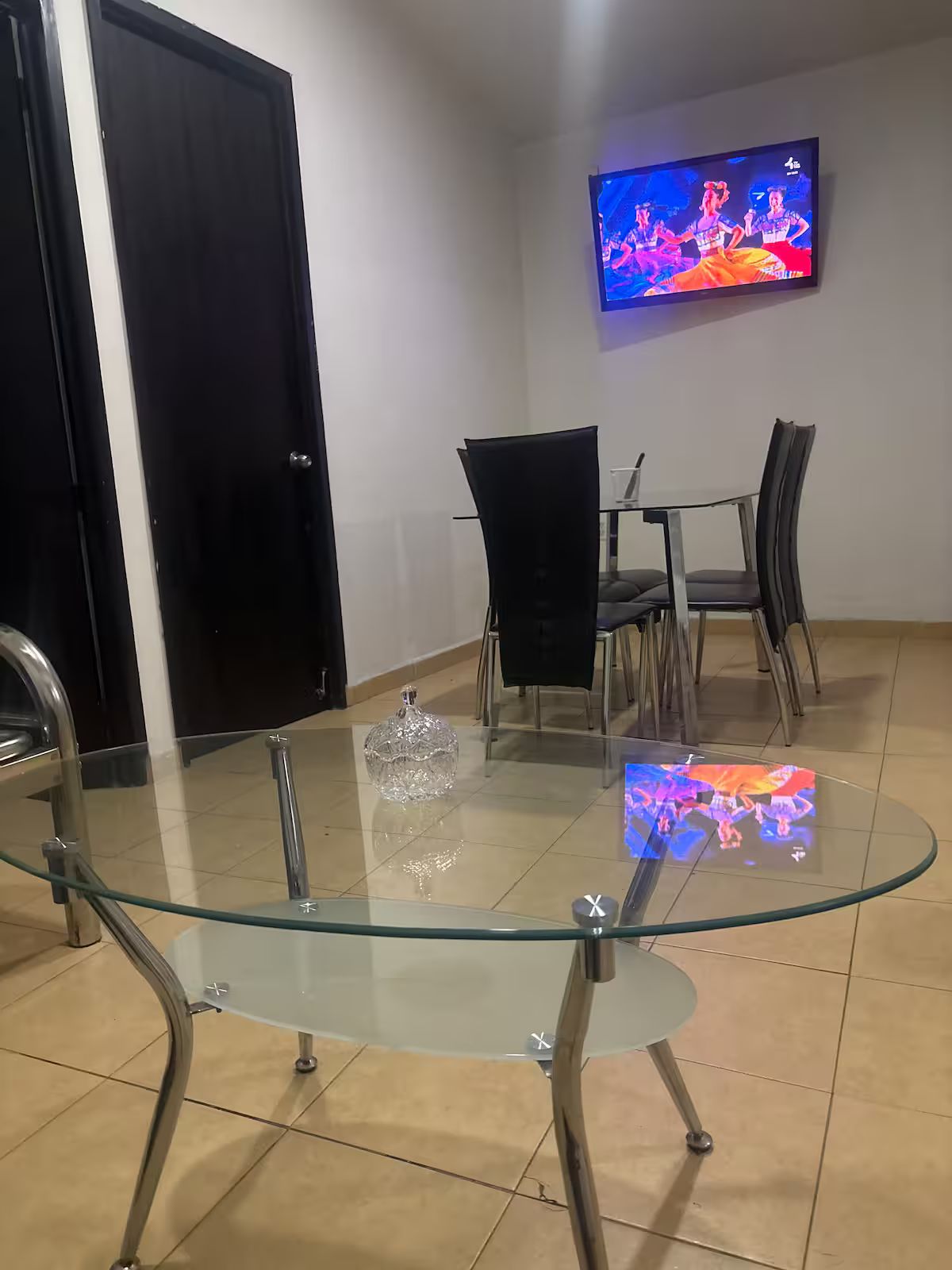 A dining area with a glass-top table, chairs, a television mounted on the wall, and a door.