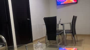 A dining area with a glass-top table, chairs, a television mounted on the wall, and a door.