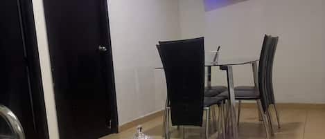 A dining area with a glass-top table, chairs, a television mounted on the wall, and a door.