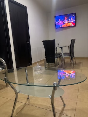 En matplats med ett bord med glasskiva, stolar, en tv som är monterad på väggen och en dörr.