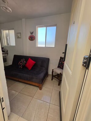 Una habitación con un sofá de color oscuro, una pequeña silla de madera, una ventana con persianas, un adorno en la pared y un piso de baldosas.
