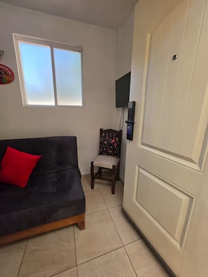A room with a dark-colored sofa, a chair with a colorful cushion, a television mounted on the wall, and a white door with a handle.