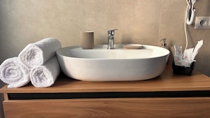 Ein Badezimmer mit einem runden Waschbecken, einem Handtuchhalter mit aufgerollten Handtüchern, einem Seifenspender und einem Taschentuchspender.