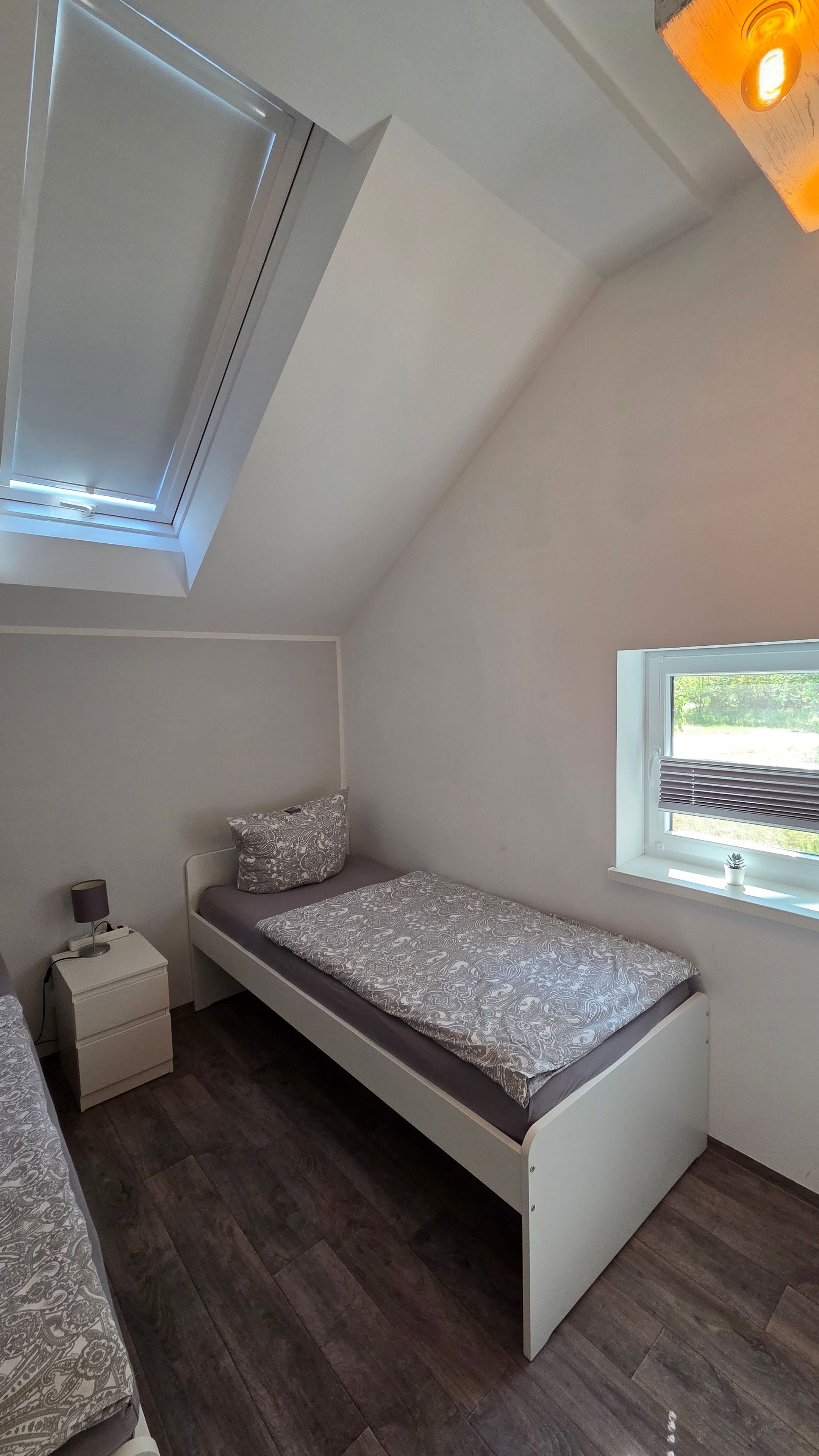 A small, single-bed room with a sloped ceiling, a window with blinds, a bedside table with a lamp, and a patterned bedspread.