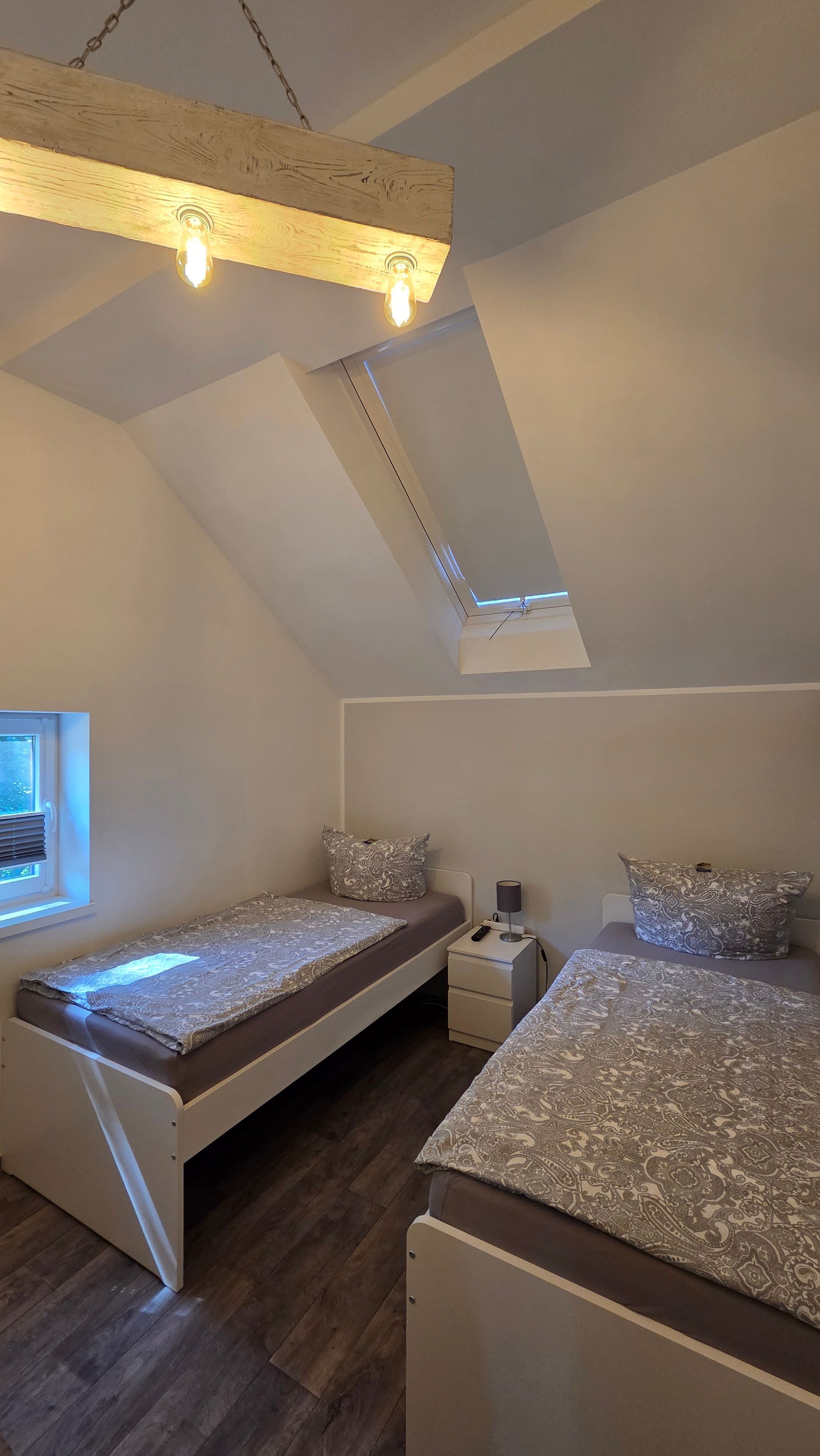 A small, well-lit attic bedroom with two beds, a window, and a skylight.