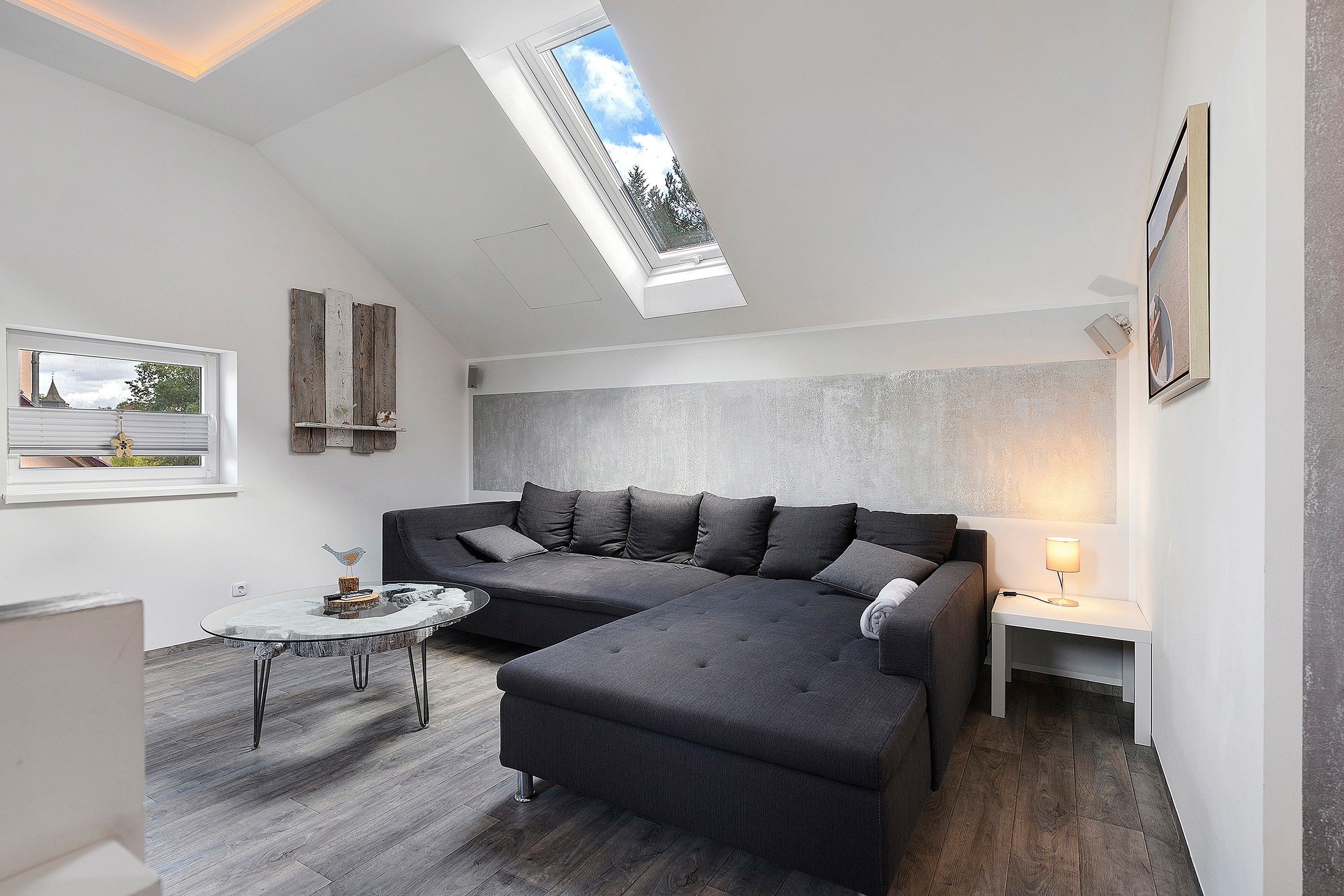 Une chambre de style loft moderne, dotée d’une lucarne, d’un canapé d’angle, d’une petite table ronde et d’une lampe de chevet.