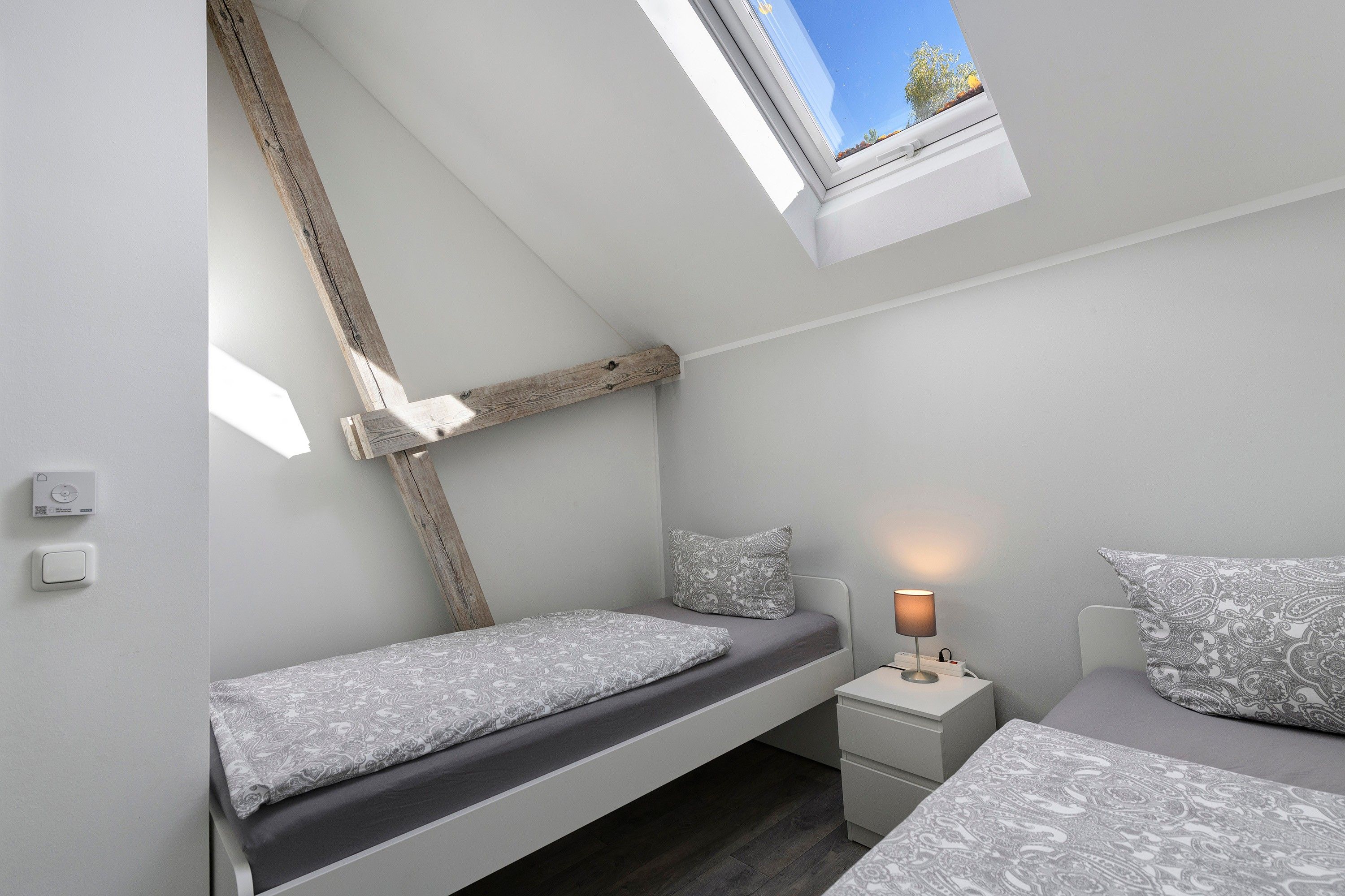 Ein Schlafzimmer mit schrägem Dach, einem Oberlicht, einem Bett mit gemusterten Bettbezügen, einem Nachttisch mit Lampe und einem an der Wand montierten Thermostat.