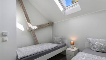 Ein Schlafzimmer mit schrägem Dach, einem Oberlicht, einem Bett mit gemusterten Bettbezügen, einem Nachttisch mit Lampe und einem an der Wand montierten Thermostat.
