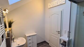 Une salle de bain avec un lavabo blanc, un miroir, un meuble de rangement, une douche avec une porte en verre et un luminaire au plafond.