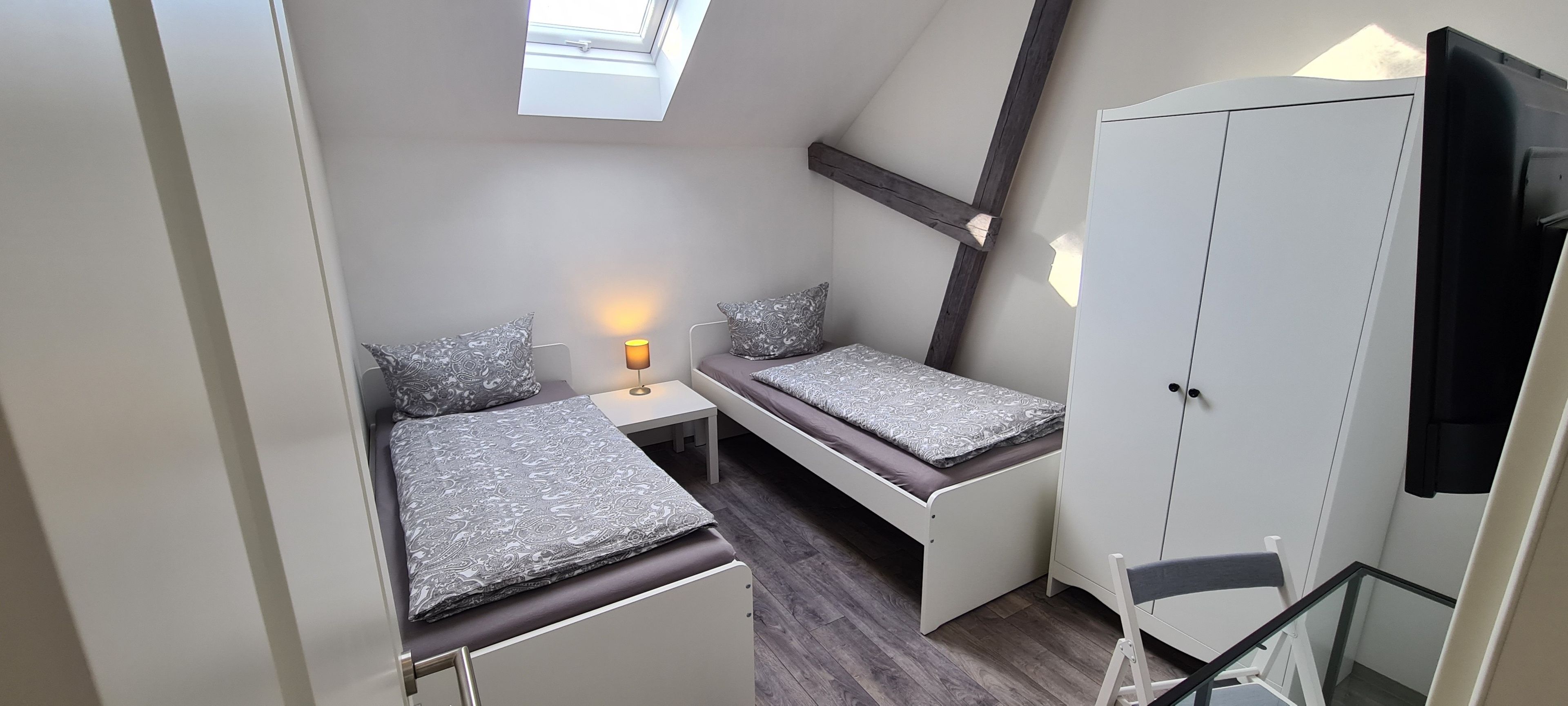 A compact room with two beds, a small table with a lamp, a wardrobe, and a window.