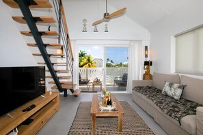 A modern living room with a TV, wooden entertainment unit, a sofa, a coffee table with a bottle, and a staircase with a ceiling fan. - Maison Sylvie  Résidence Maracuja Orient Bay (Orient bay)