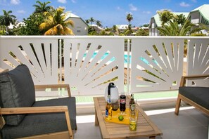A balcony with a white decorative fence, a wooden table with drinks, and a view of a pool and palm trees. - Maison Sylvie  Résidence Maracuja Orient Bay (Orient bay)