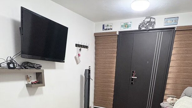 Een slaapkamer met een bed, een aan de muur bevestigde televisie, een kledingkast en een klein plankje met spullen.