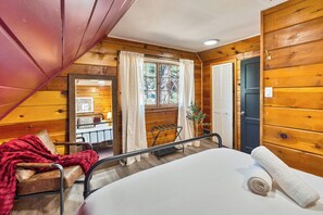 Ein gemütliches, blockhausähnliches Zimmer mit einem Bett, einem Sessel, einem kleinen Tisch und einem Fenster mit Vorhängen.