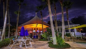 Un área de comedor al aire libre con mesas y sillas, un espacio de estar techado y una piscina al fondo.