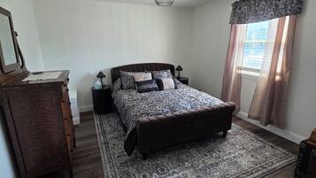 A bedroom with a large bed, a wooden dresser, a nightstand, a floor lamp, and a ceiling light fixture.