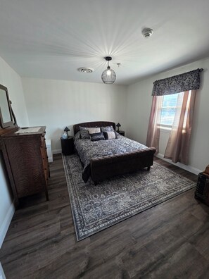 A bedroom with a large bed, a wooden dresser, a nightstand, a floor lamp, and a ceiling light fixture.
