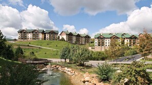 Un hotel de varios pisos con techo verde, ubicado en una ladera con un pequeño estanque y un puente de madera.