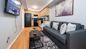 Uma sala de estar moderna com um sofá cinza, uma televisão de tela plana na parede e uma cozinha ao fundo.
