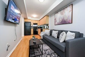 A modern living room with a grey sofa, a flat-screen TV mounted on the wall, and a kitchen in the background. - Comfort Townhouse Suites (Mississauga)