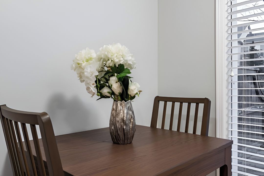 A dining table with a vase of white flowers, two wooden chairs, and a window with blinds.