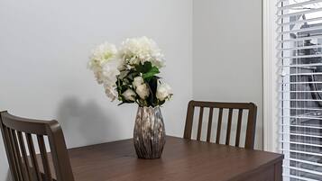 A dining table with a vase of white flowers, two wooden chairs, and a window with blinds.