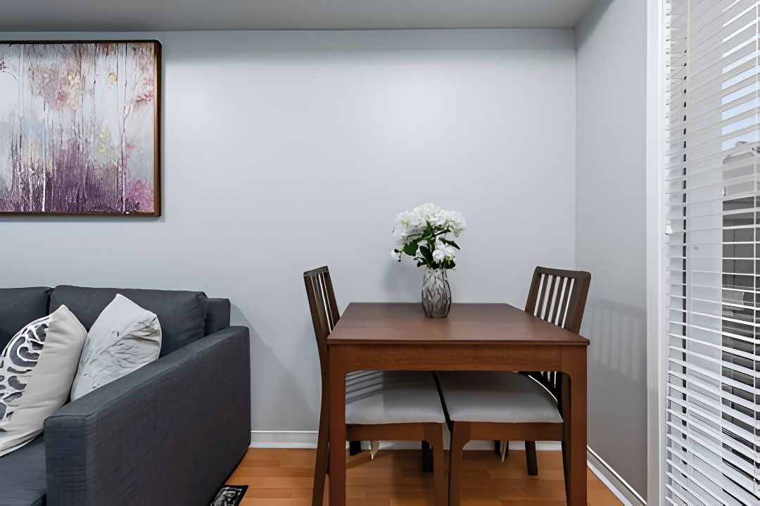 A dining area with a wooden table and chairs, a vase with white flowers, and a grey sofa with patterned cushions.