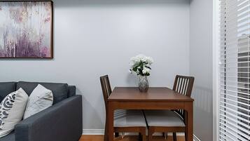 A dining area with a wooden table and chairs, a vase with white flowers, and a grey sofa with patterned cushions.