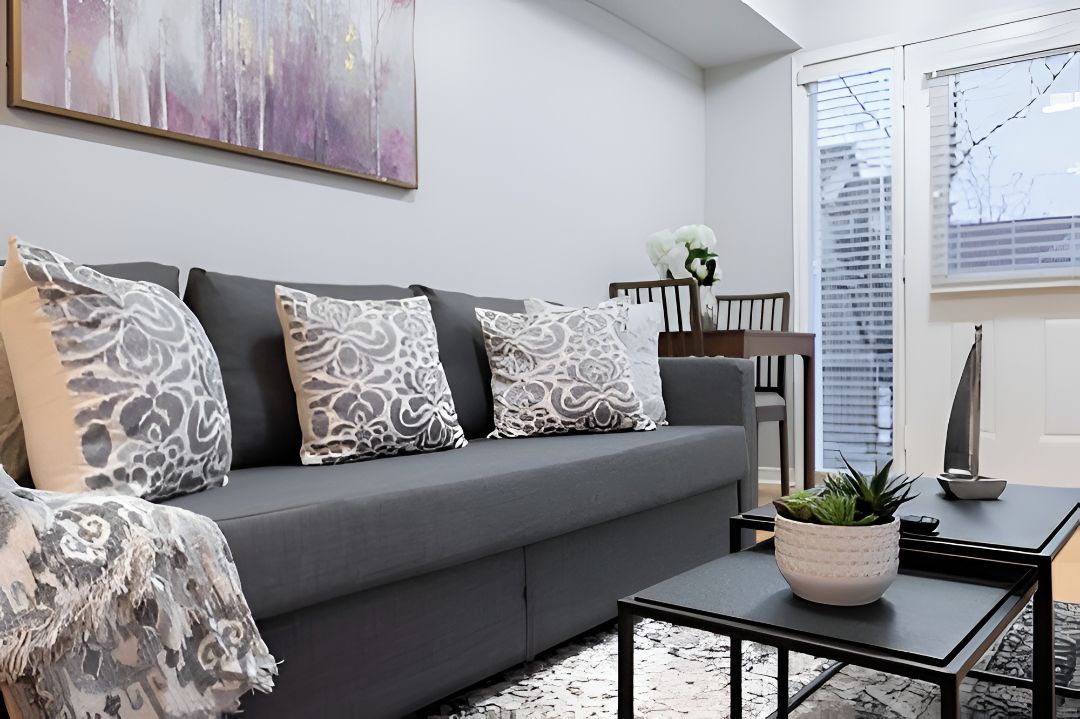 Un salon moderne avec un canapé gris, des coussins à motifs, une table basse agrémentée d’une plante et un tableau accroché au mur.