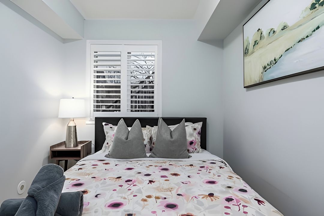 A bedroom with a bed, a nightstand, a lamp, a chair, and a window with blinds.