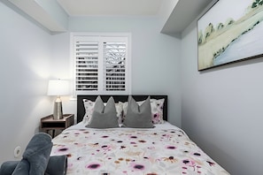 A bedroom with a bed, a nightstand, a lamp, a chair, and a window with blinds.