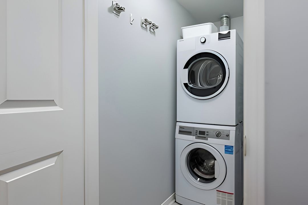 A compact laundry room with a stacked washer and dryer, a white door, and a wall-mounted towel dispenser.