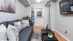 Uma sala de estar moderna com um sofá cinzento, almofadas estampadas, uma mesa de centro preta com uma planta, um quadro abstrato grande e uma TV montada exibindo diversos canais.