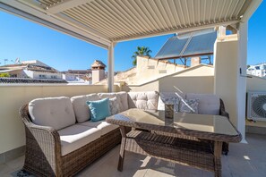 A covered outdoor seating area with a wicker sofa and table, solar panels, and a view of buildings in the background. - Holiday Home in Nerja With Sea Views (Nerja)