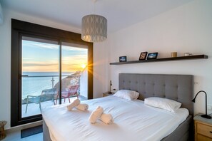 A hotel room with a large bed, a view of the sea, a balcony with chairs, and a shelf with framed photos. - Apartment in Nerja With Panoramic Sea Views (Nerja)