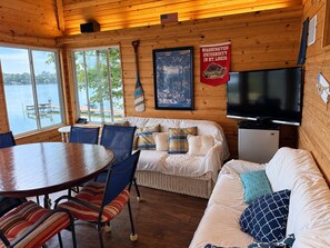 A cabin with a dining area, a sofa, a TV, and a poster of a sports event. - Beautiful Gun Lake Cottage – Private Lakefront, Sandy Bottom & Lots of Lake Toys (Middleville)