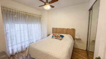 Un dormitorio con una cama, un ventilador de techo, una ventana con cortinas translúcidas y suelo de madera.