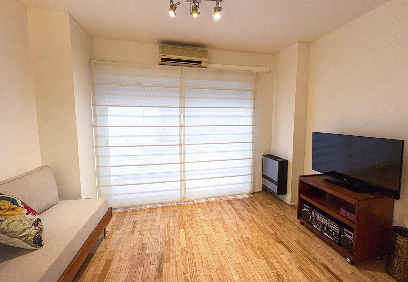 A living room with a sofa, a television on a wooden stand, and a window with blinds.