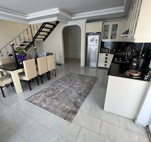 Eine moderne Küche mit Kühlschrank, Herd und Schränken. Es gibt einen Essbereich mit Tisch und Stühlen. Im Hintergrund ist eine Treppe zu sehen.