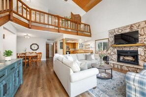 A spacious living area with a high ceiling, a fireplace, a television mounted on a stone wall, and a dining area with wooden furniture. - Coastal Retreat Big Sur Style • King Beds • EV+Pet (Cambria)