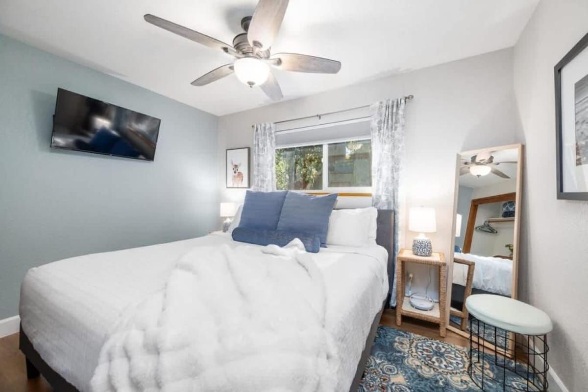 A bedroom with a bed, a ceiling fan, a window with curtains, a bedside table with a lamp, a mirror, and a patterned rug.