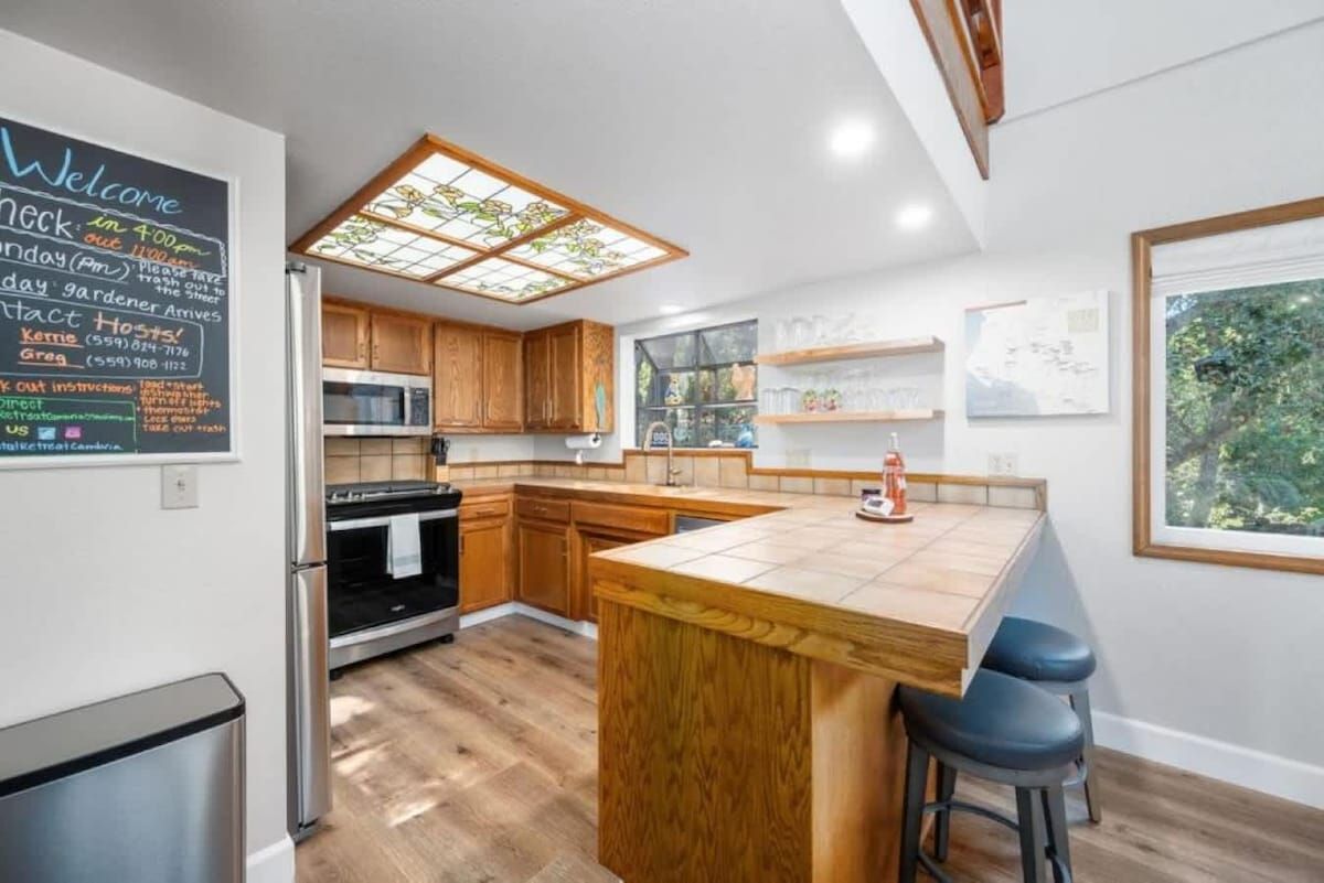 A modern kitchen with wooden cabinets, a central island with stools, a chalkboard with contact information, and a large window with a view of greenery.