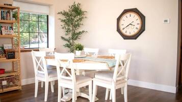 A dining area with a wooden table, white chairs, a clock on the wall, and a bookshelf.