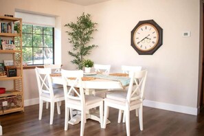 A dining area with a wooden table, white chairs, a clock on the wall, and a bookshelf. - Coastal Retreat Big Sur Style • King Beds • EV+Pet (Cambria)