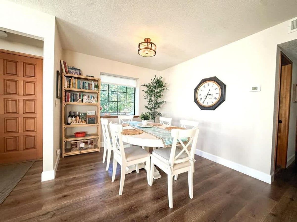 A dining area with a wooden table and chairs, a bookshelf, a clock on the wall, and a door leading to another room.