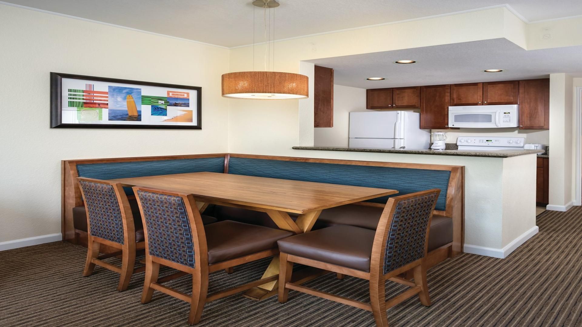 A hotel room with a dining area featuring a wooden table and chairs, a kitchen with a refrigerator and microwave, and a framed picture on the wall.