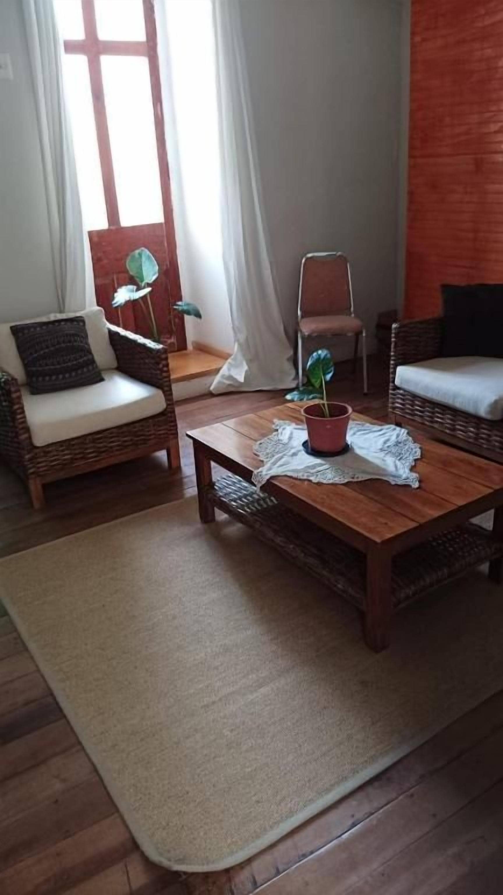 Una habitación con dos sillas de mimbre, una mesa de centro de madera, una planta en maceta y vistas a través de una ventana con cortinas.